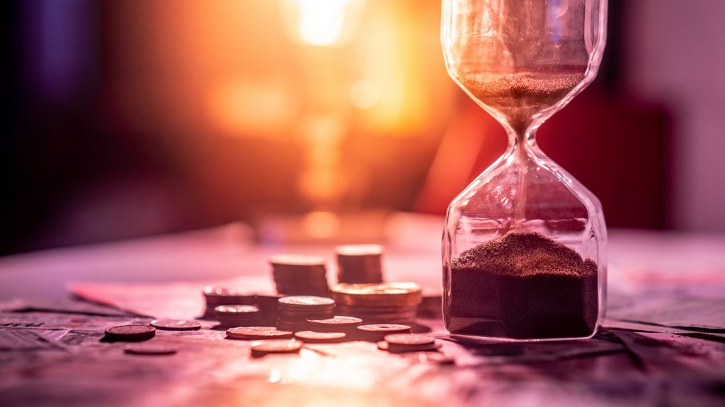 Sand running through the shape of hourglass on table with banknotes and coins of international currency. Time investment and retirement saving. Urgency countdown timer for business deadline concept